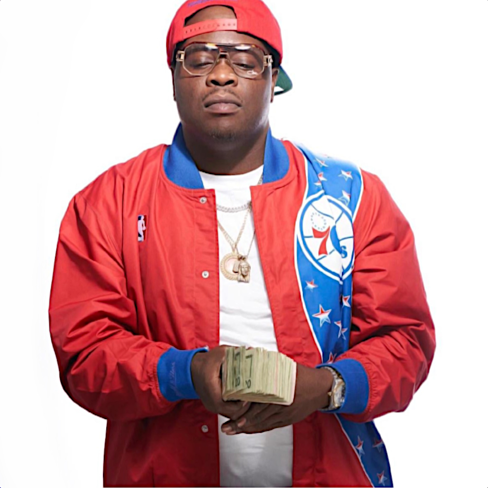 Trong hình, rapper người Mỹ E Ness đứng vững chãi, hướng về phía trước, đầu ngẩng cao, đội một chiếc mũ bóng chày màu đỏ ngược và mặc một chiếc áo khoác đỏ đồng bộ; hai tay anh ấy đầy những tờ tiền mệnh giá 100 đô la. Toàn bộ khung cảnh tràn ngập sắc màu rực rỡ và năng lượng sôi động.
