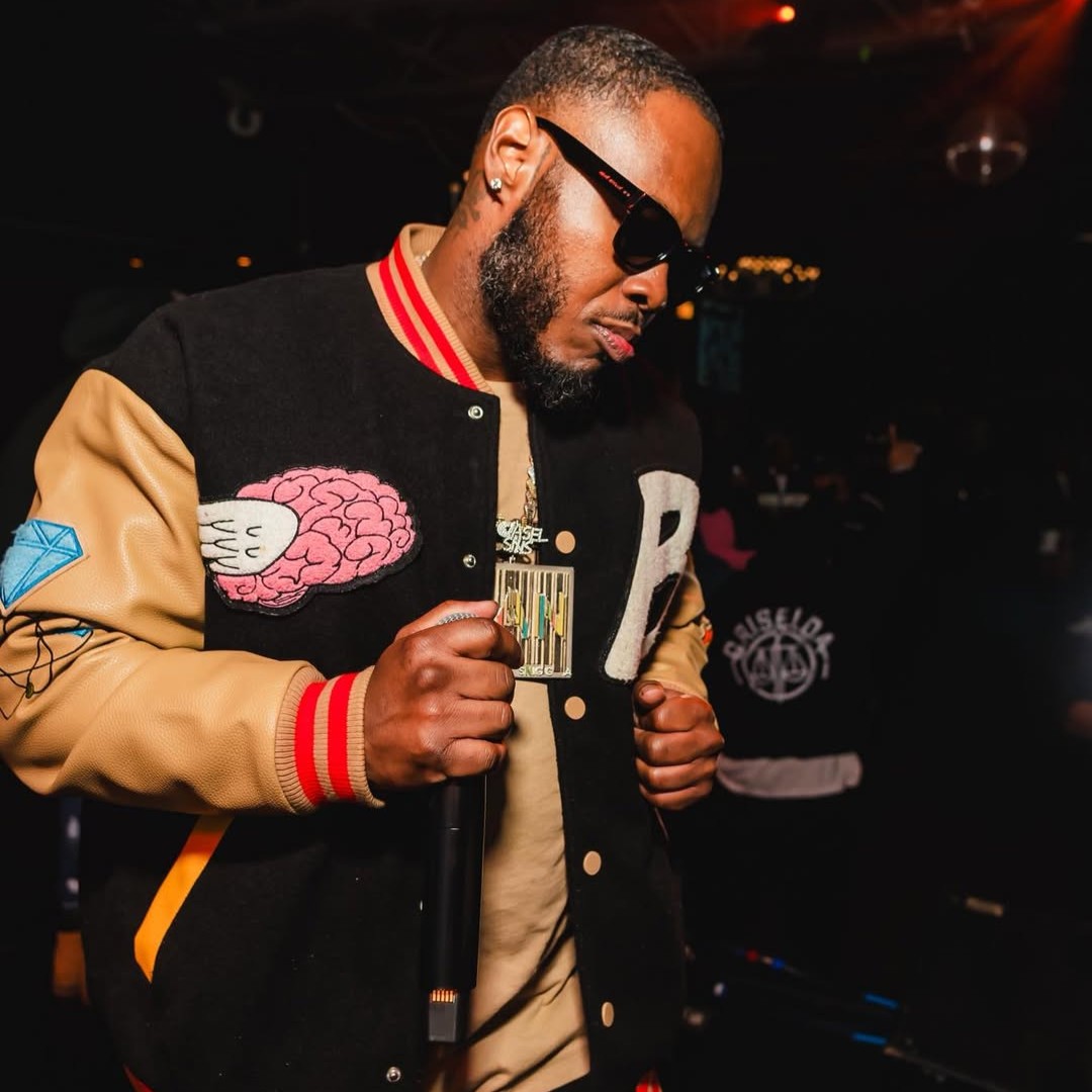 In the image, American rapper Rufus Sims stands facing left, head slightly lowered, wearing sunglasses and a baseball jacket with leather sleeves. His right hand holds a microphone, while his left sits bent at his side. The whole scene bursts with bold colors and vibrant energy.