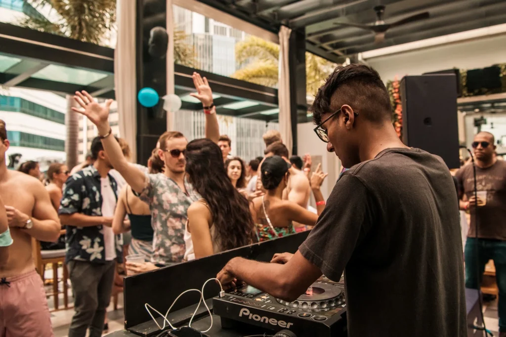 Singapore-based DJ Sivanesh performs behind a Pioneer DJ setup in an open bar area, playing to a daytime crowd during a house-focused set.