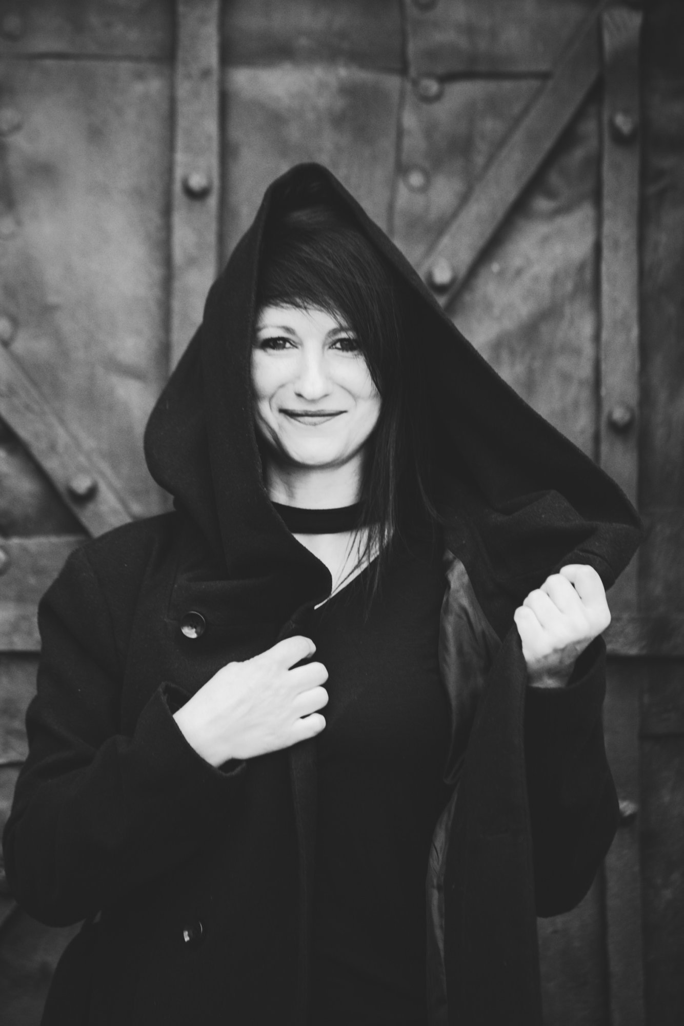 Slovenian Female DJ Ani Onix standing outside gate wearing a hooded coat clasping the hood with left hand whilst smiling, black and white image.