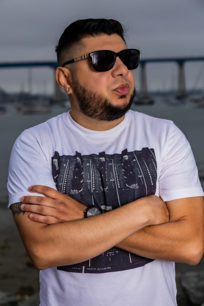 American DJ and radio presenter DJ Nocturne is portrayed in a lively scene by the harbor, gazing to his left with arms crossed. He sports a white t-shirt emblazoned with a Pioneer DJM800 mixer design and a pair of sunglasses, set against the bustling