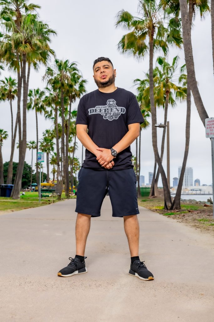 American DJ and radio host DJ Nocturne is captured in a vibrant upright shot, gazing at the camera with a playful smirk. He sports a DJ Nocturne baseball cap and an “On the Deep End” T-shirt, standing with both arms in front and his hands clasped tog