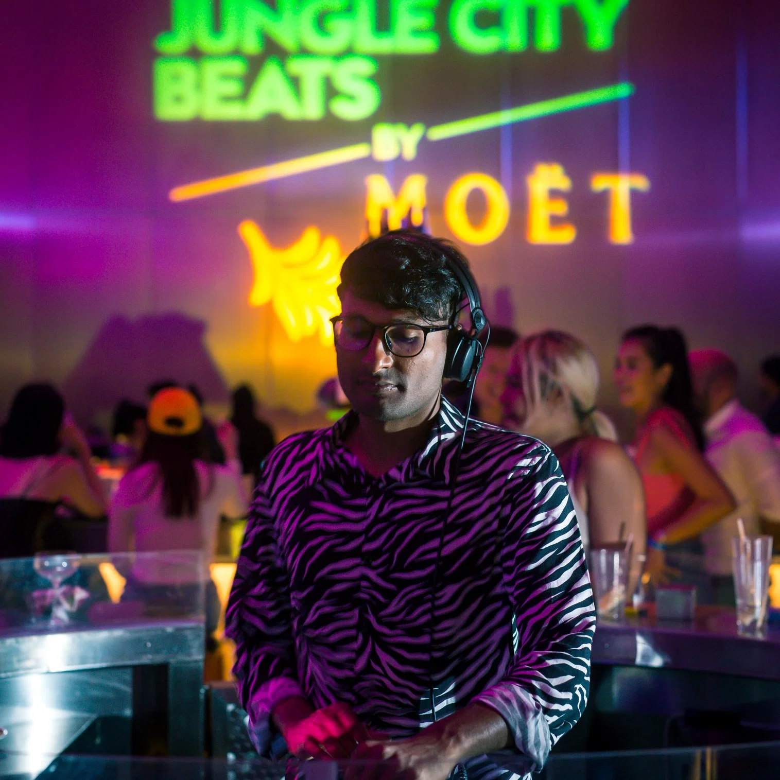 Singapore-based DJ Sivanesh performs at Jungle City Beats, wearing headphones in front of a club crowd with colourful event visuals behind him.