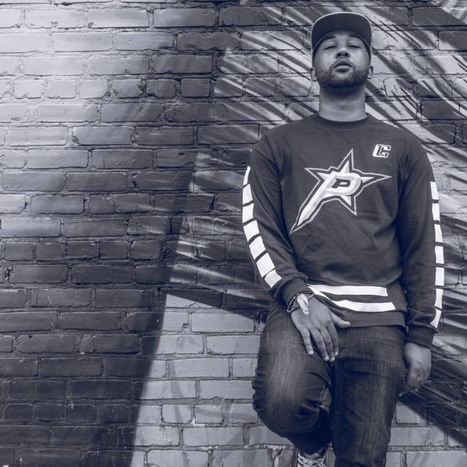 Black and white photo of underground hip hop artist Bhramabull standing against a brick wall with graffiti, wearing a cap and casual streetwear.