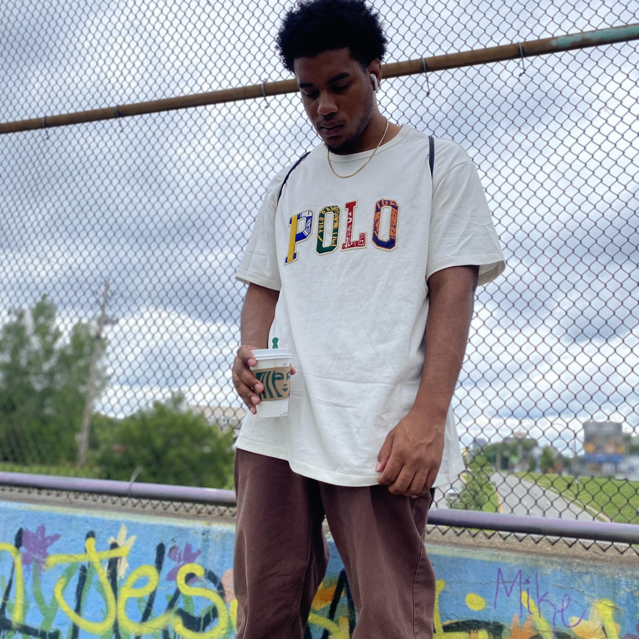 West Coast rapper Kail Problems standing outdoors near a chain-link fence, wearing a Polo T-shirt and holding a coffee, captured in a casual urban setting.