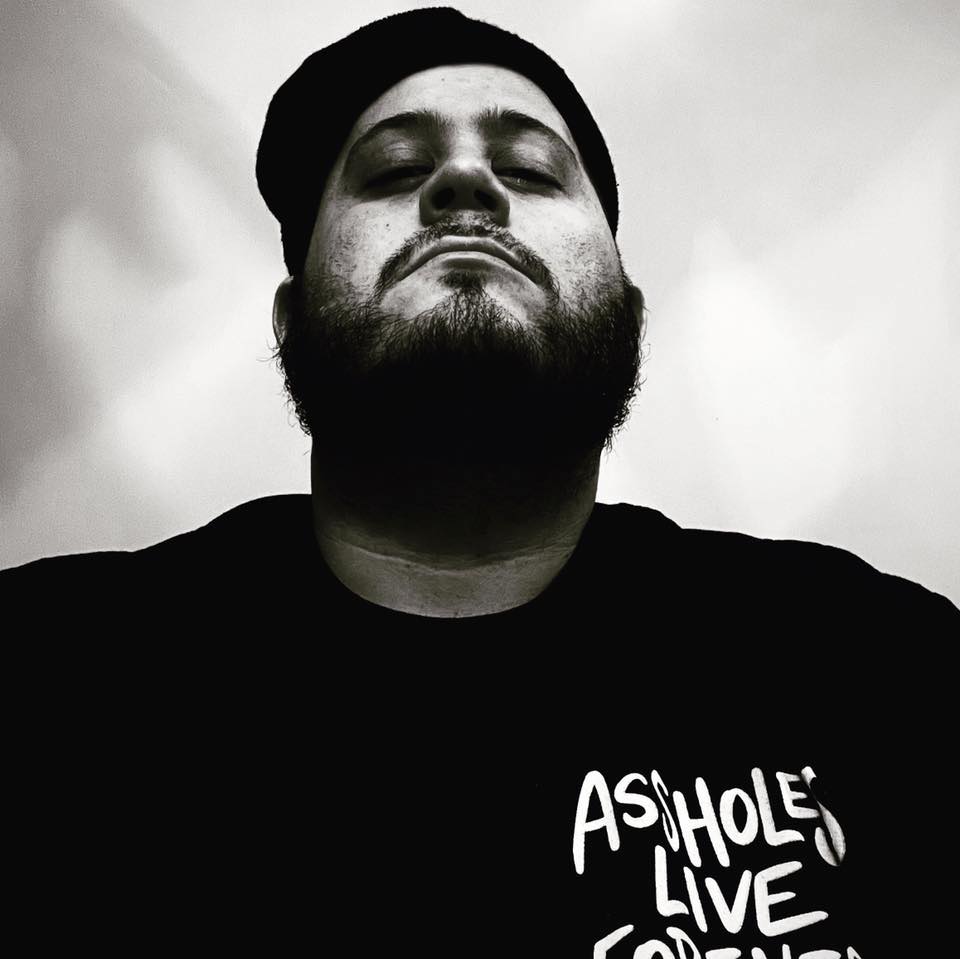 Black and white portrait of underground hip hop artist Coast Locastro, wearing a cap and T-shirt, photographed from a low angle with a bold, confident expression.