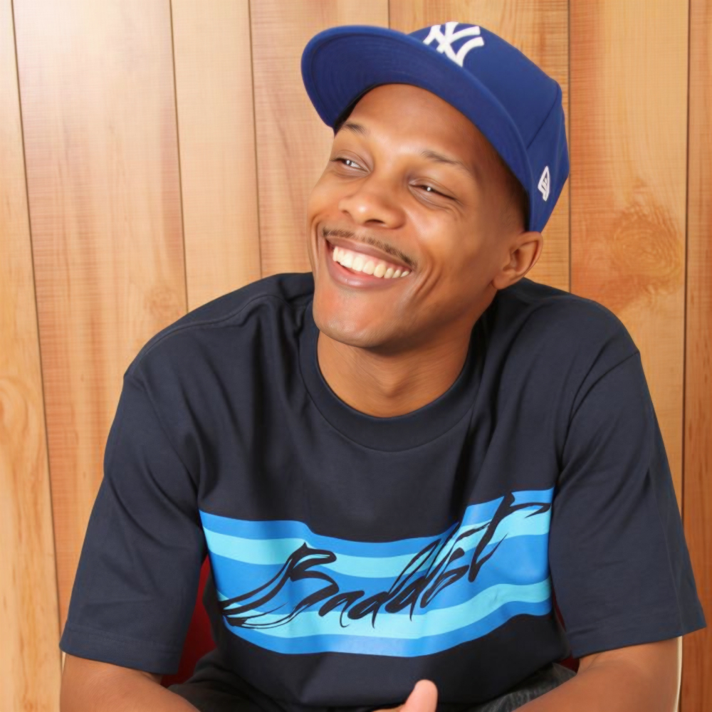 Portrait of Wordsworth, a New York-based hip hop artist, smiling and wearing a blue cap and T-shirt, seated against a wooden background.