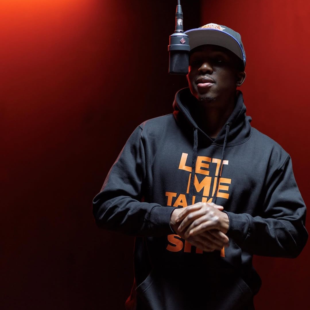 Portrait of Rigs, hip hop artist, wearing a cap and hoodie, standing under a microphone against a red background with hands clasped.