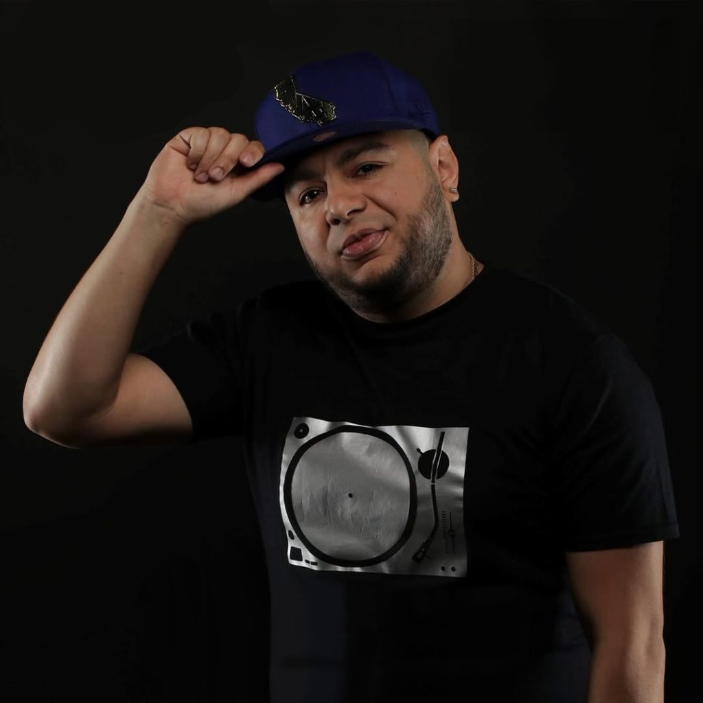 Portrait of DJ Nocturne, host of Cali’s HouseNation Radio, wearing a cap and black T-shirt while holding the brim of his hat against a black background.