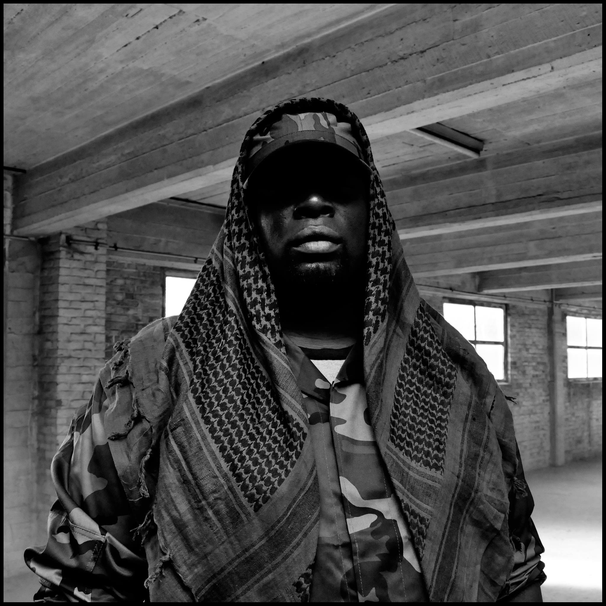 Black and white portrait of Chip-Fu wearing a cap and patterned Shemagh scarf, standing in an industrial indoor setting with a shadowed face.