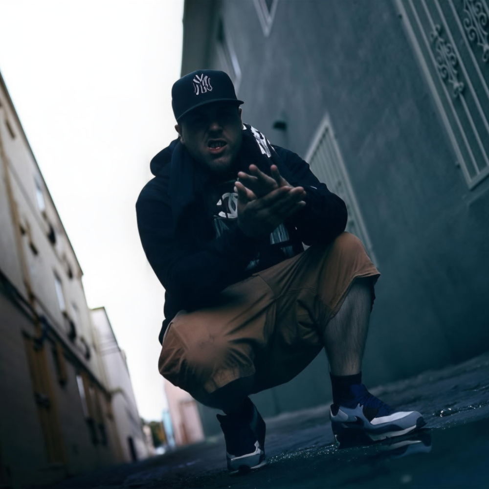 Johnny Slash crouching in an urban alleyway, wearing a cap, hoodie and shorts, with a low-angle perspective and moody lighting creating a gritty street atmosphere.