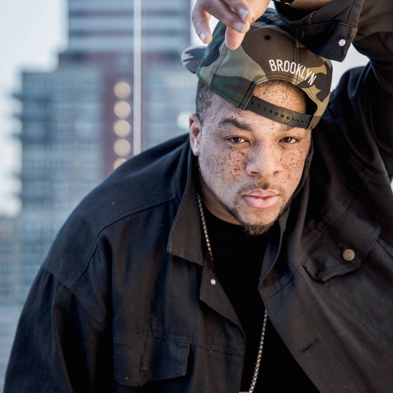 Brooklyn hip hop artist Mic Handz posing in a black jacket and camouflage Brooklyn cap against a blurred city skyline background.