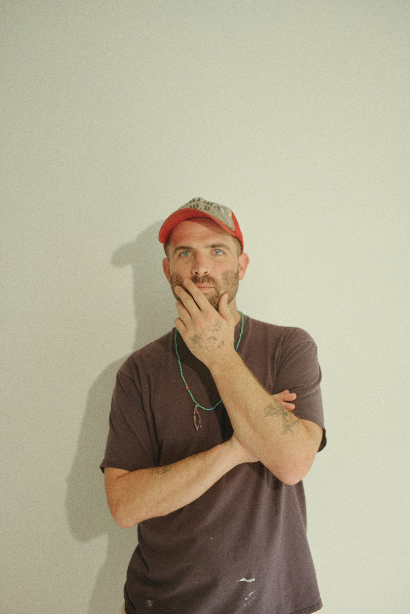 Portrait of designer and creative collaborator Sean Wotherspoon standing against a minimalist white wall, wearing a red cap and casual dark T-shirt. With folded arms and a thoughtful expression, the image reflects his multidisciplinary approach to design, sustainability and contemporary culture.