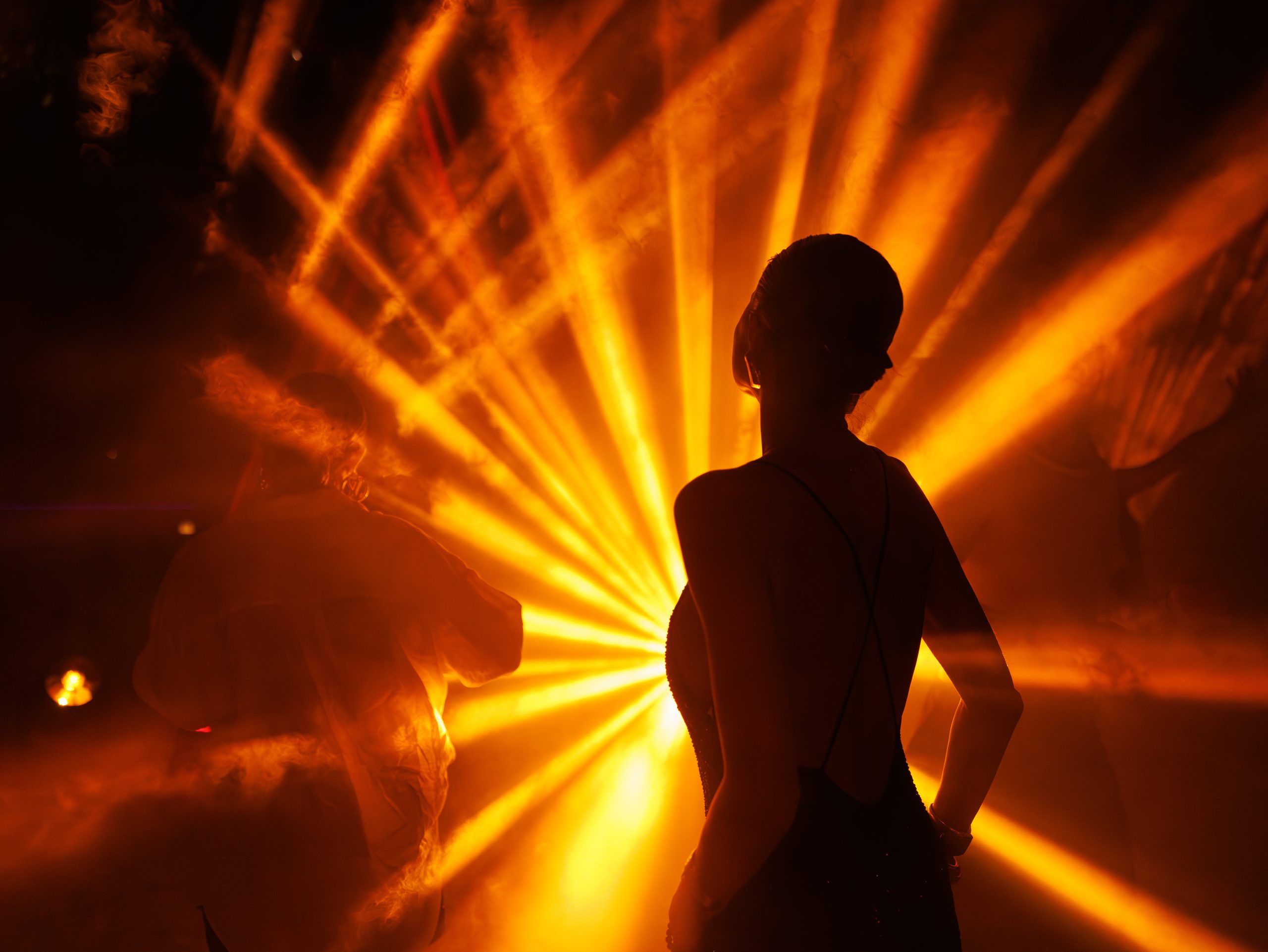 Guests gather beneath dramatic golden stage lighting during a previous Fari Islands Festival event. Silhouetted figures stand and socialise amid atmospheric haze as powerful beams of light radiate across the venue, creating an immersive nightlife experience.