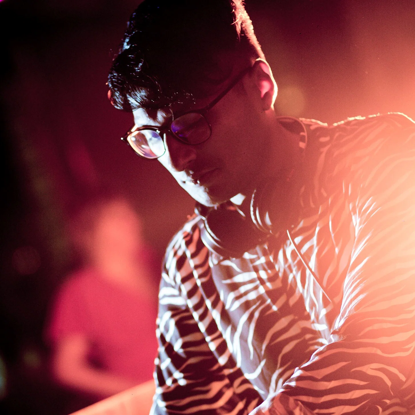 Close-up image of Singapore-based DJ Sivanesh wearing glasses and headphones around his neck, lit by warm red club lighting during a house and groove-driven techno set.