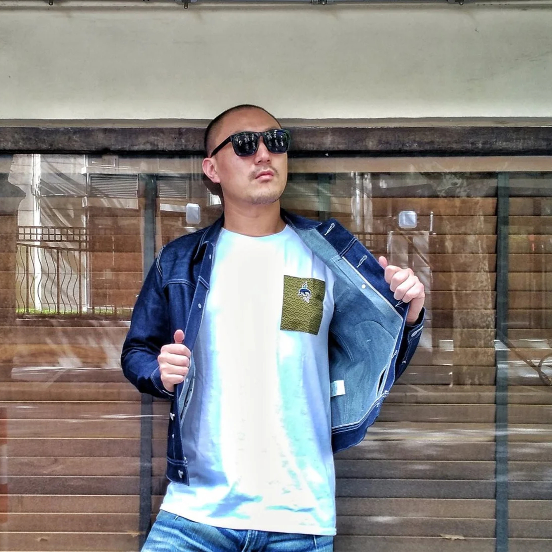 Murphy Kin in sunglasses opens a denim jacket over a white graphic T-shirt, posing confidently in front of a window with blinds.