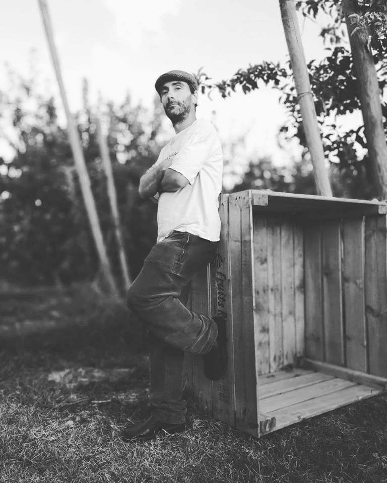 Spanish DJ and Producer A503X leaning agaist large wooden box with arms crossed wearing a hat, black and white image.
