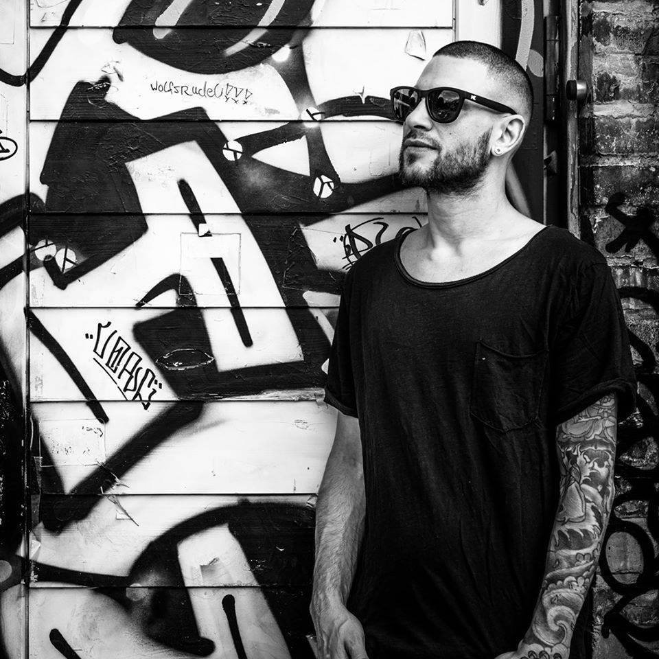 Argentianian DJ and producer Pablo Pitch standing by b=graffiti wall wearing sunglasses 45 degrees to right.