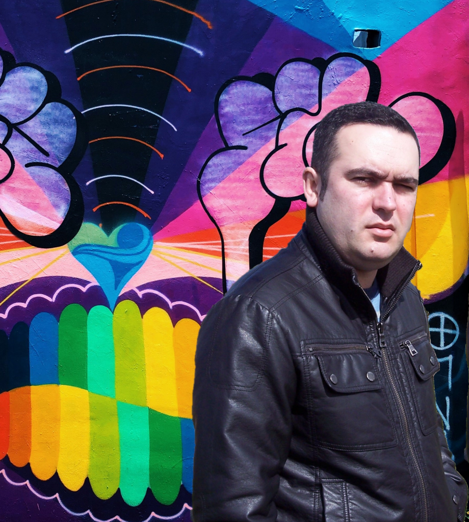 Portuguese Dj Joshua stading in front of a mural facing left wearing a leather jacket, colour image.
