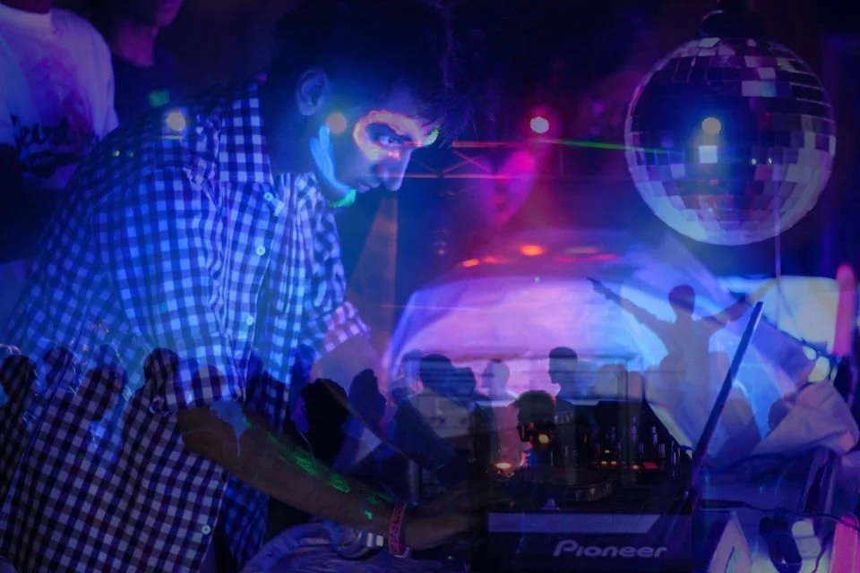 Pakistani DJ and Producer DJ Mawji performing on Pioneer CDJs at an event, side shot, colour image.