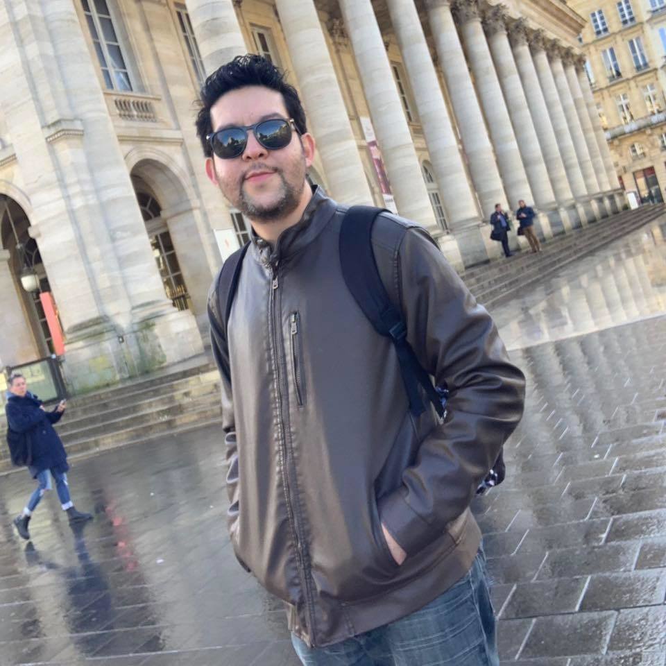 American D Diaga standing in front of building with hands in jacket pockets wearing sunglasses, colour image.