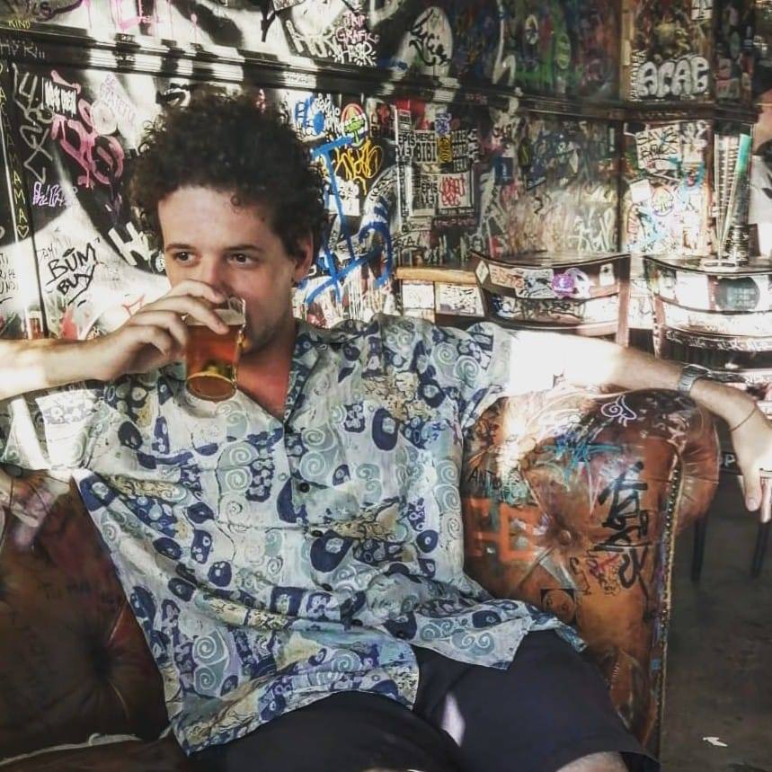 Italian Techno Dj pulsɘs sipping on a drink whilst sitting in a chair, colour image.
