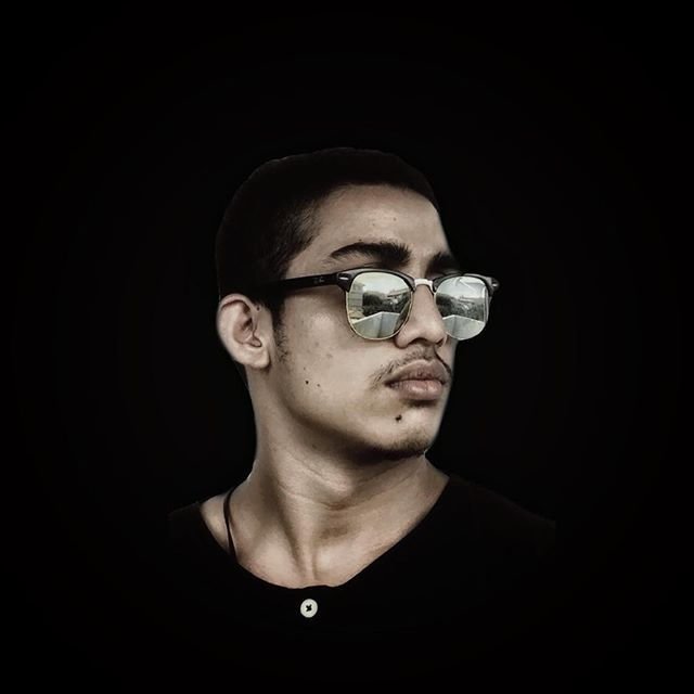 Sri Lankan DJ Deep A looking left wearing sunglasses against a black background, colour image.
