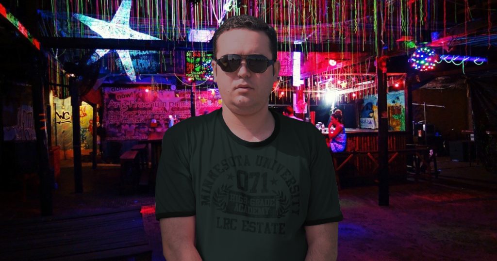 Portuguese DJ Joshua standing in a open bar wearing sunglasses after sundown, colour image.
