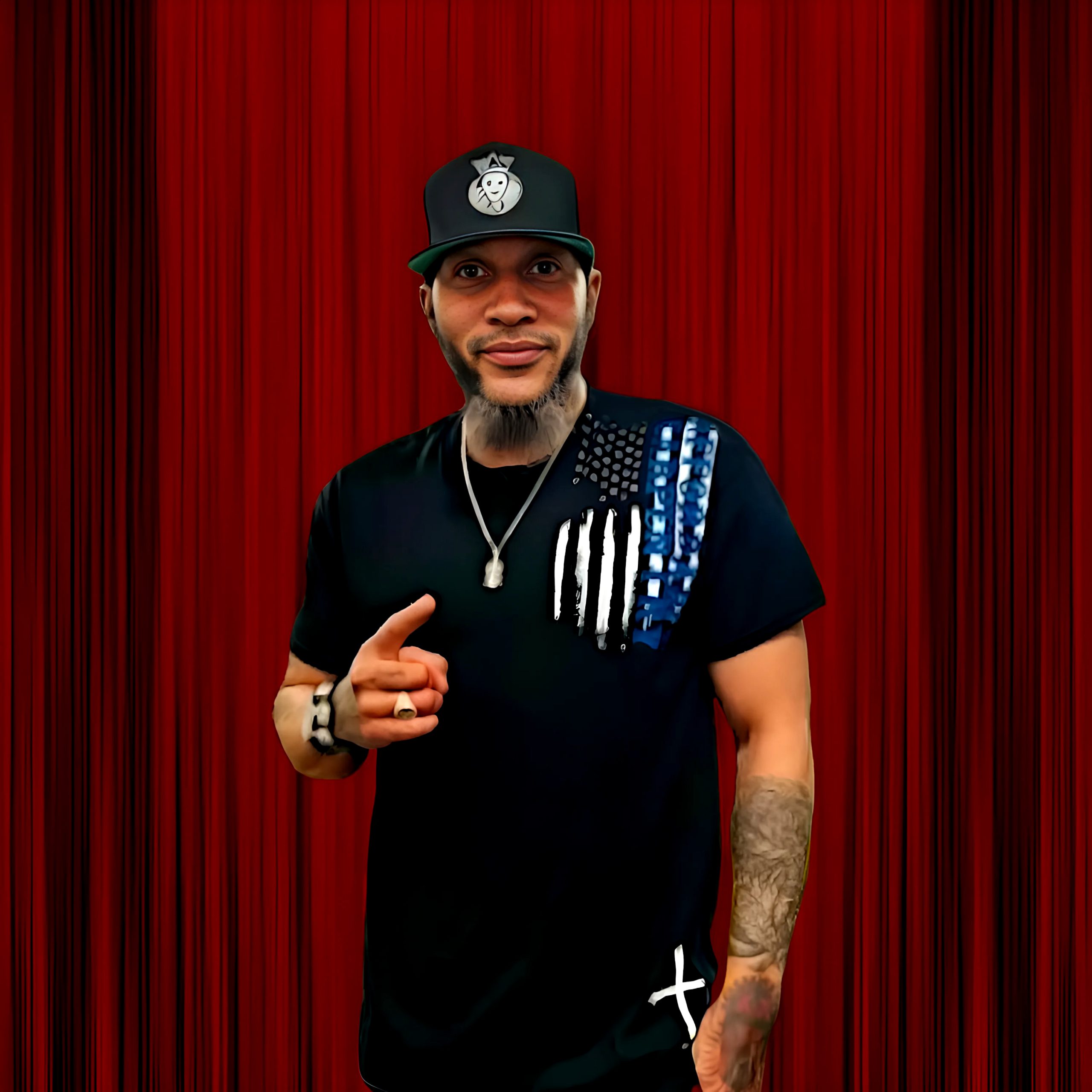 American Rapper and producer Krumbsnatcha standing in front of a red curtain with his right finger pointing forward and his left hand in his pocket wearing a baseball cap, colour image.
