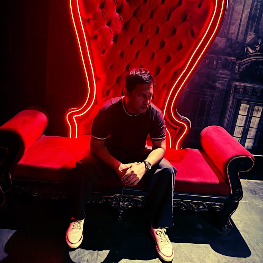 Indian DJ Kafka sitting on a plush armchair covered in red velvet with a huge back and arm rests looking to his left with his hands on top of each other, colour image.