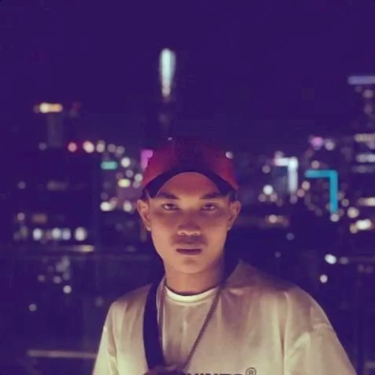 Vietnamese DJ Tommy wearing a Baseball Cap standing in front of a high window looking out on Ho Chi Minh City Night Skyline, colour image.