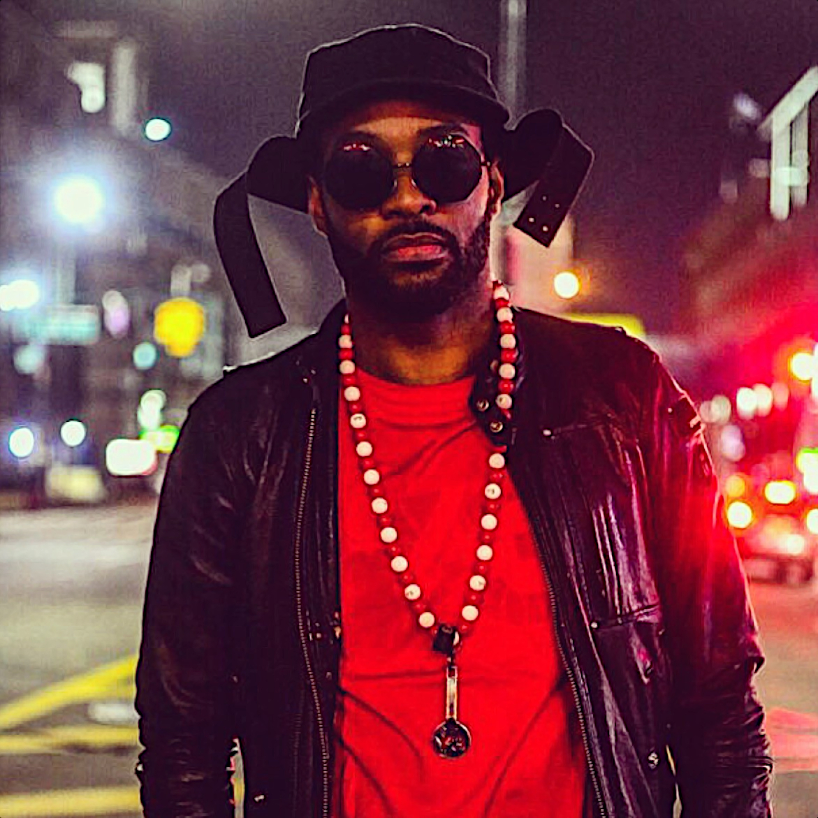 American rapper Mickey Factz walking down the street wearing sunglasses and a leather jacket over a red t shirt with a hat at night time, colour image.