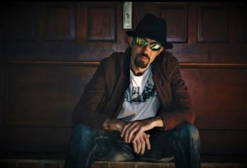 New Zealand DJ Snuck sitting outside a door on a step wearing sunglasses and a hat.