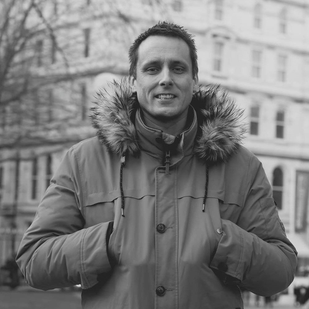British DJ and producer Kev Willis stading in a warm coat with hands in pockets smiling, black and white image.