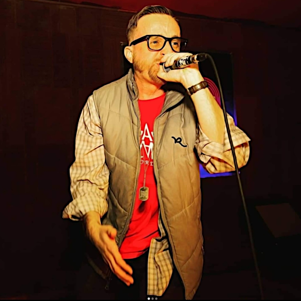 Rapper, nhà sản xuất và kỹ sư người Mỹ Jake Palumbo biểu diễn tại FM Lounge ở Jersey City, NJ, Hoa Kỳ, đeo kính và mặc áo vest, tay trái cầm micro đưa lên miệng, ảnh màu.