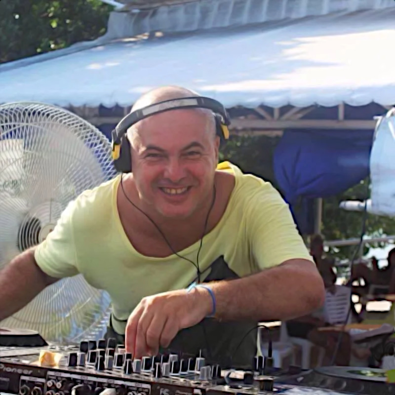 Italian DJ and producer Marco Mei performing at an outdoor event leaning forward over the DJ decks and smiling, colour image.