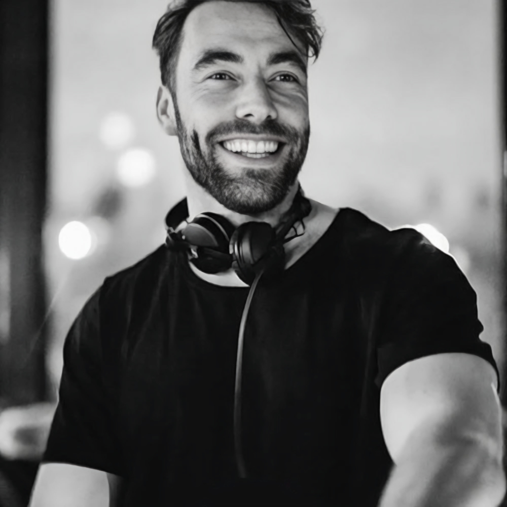 Black-and-white portrait of Amsterdam DJ Jack Delhi smiling with headphones around his neck, representing his House music background and work with The Wild Bunch.