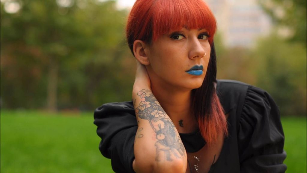 Female Romanian Techno DJ Deysa holding back of neck looking at camera, wearing blue lipstick.