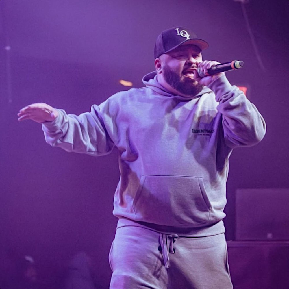 American rapper Tony Moxberg performing at an event wearing a baseball cap and hoodie tracksuit holding the microphone with his left hand in square colour image.