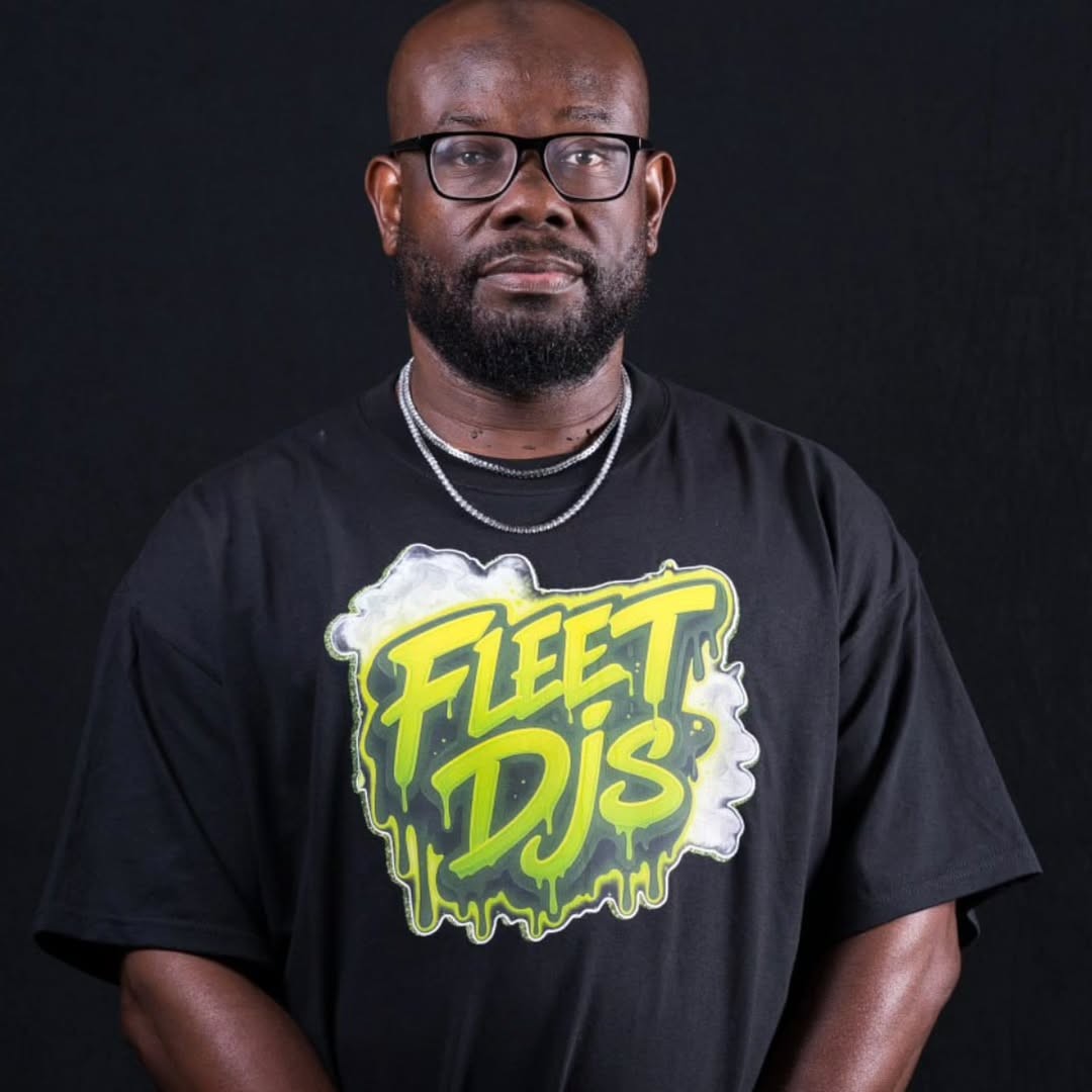 Trong bức ảnh, nhà sản xuất người Mỹ, CEO của Fleet DJs và rapper KLASSIK đang mặc một chiếc áo phông Fleet DJs, đeo kính và quần jean; anh ấy đang đứng nhìn thẳng vào máy ảnh; hình ảnh được chụp màu.