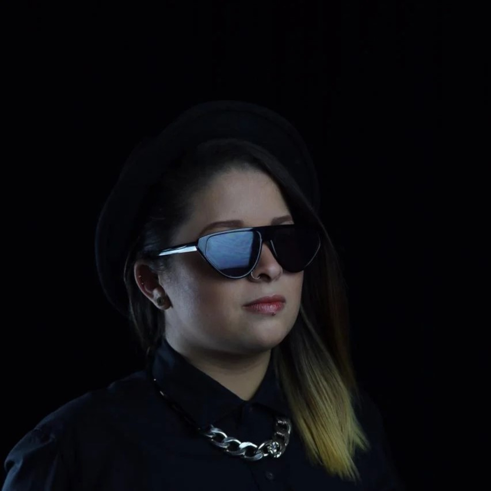 Venezuelan DJ and Producer Natasha Delgado standing in dark room wearing sungasses and a hat looking to the left.