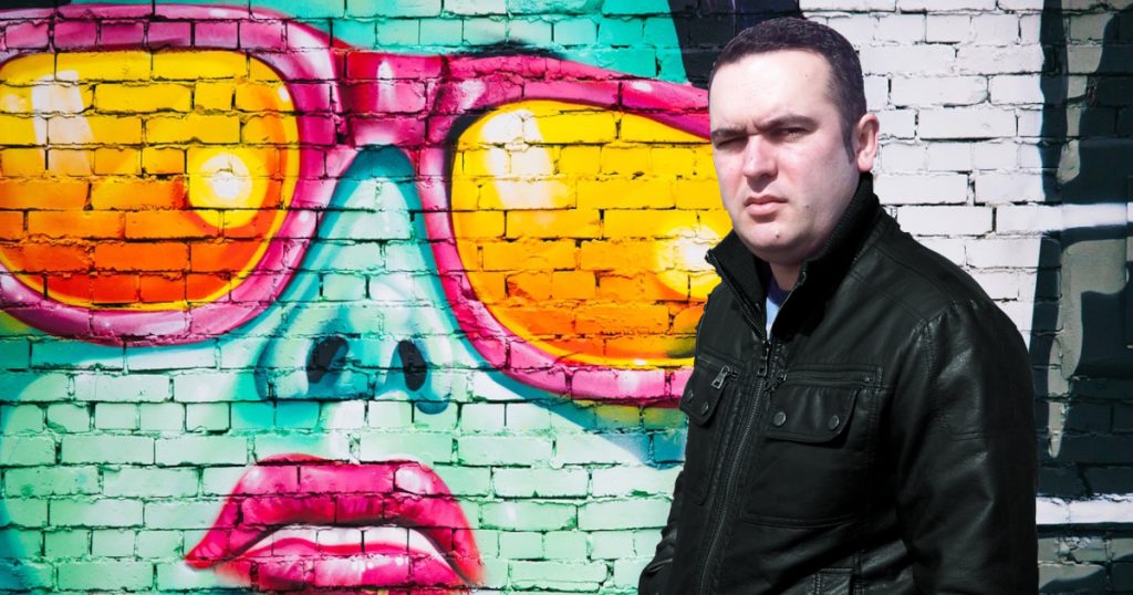 Portuguese DJ Joshua standing in front of Graffiti with a woman wearing sunglasses, colour image.