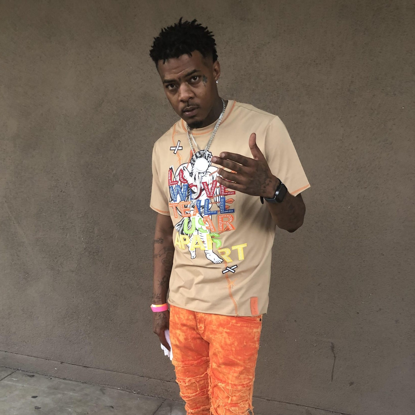 Nhà sản xuất, rapper và chủ hãng thu âm người Mỹ Junya Boy đứng với tay trái giơ lên, cánh tay phải để bên hông, mặc quần jean denim màu cam và áo phông màu be, nhìn thẳng vào ống kính trong một bức ảnh màu.