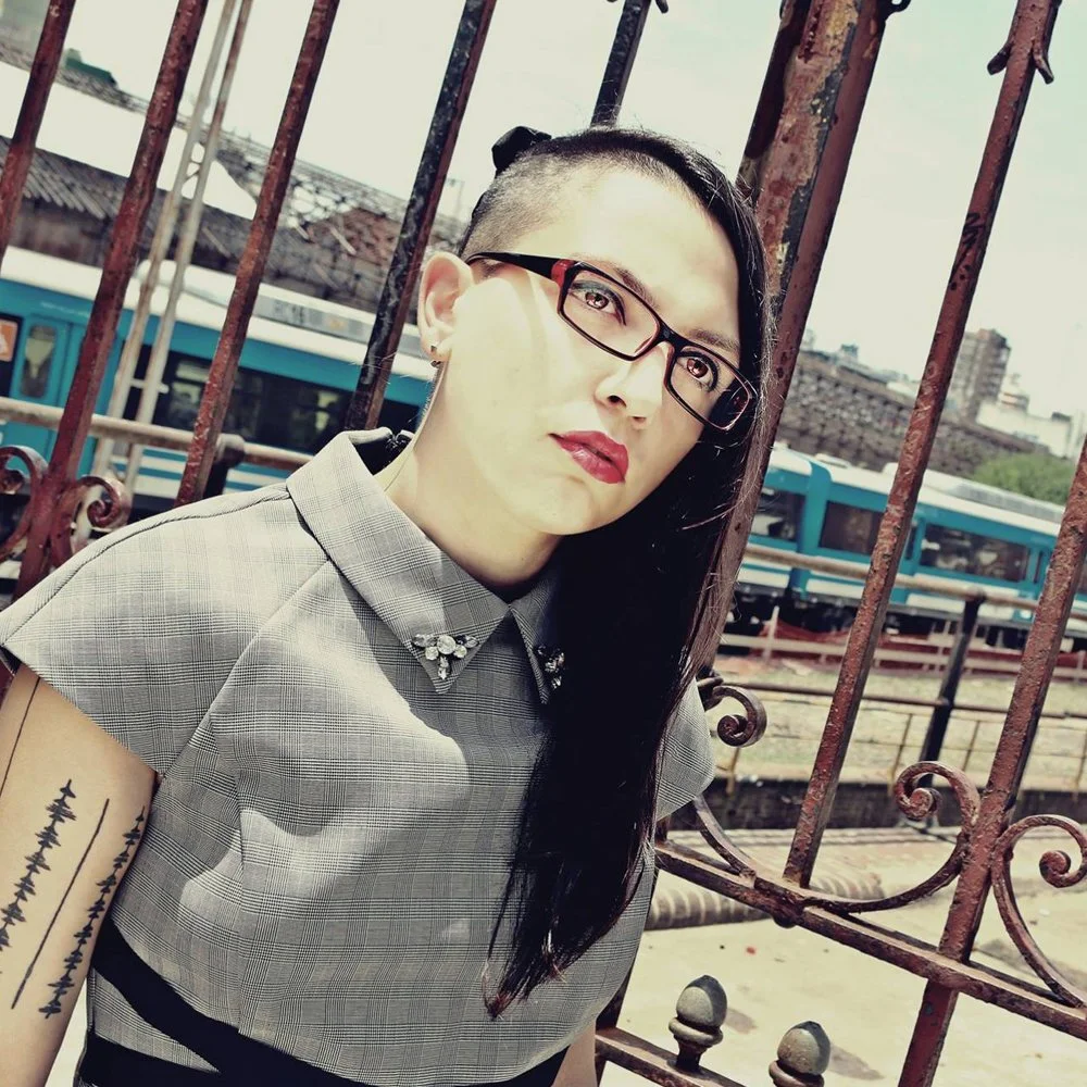 Argentinian Female DJ and producer Miss Adk standing beside a gate outside a train track wearing glasses, colour image.