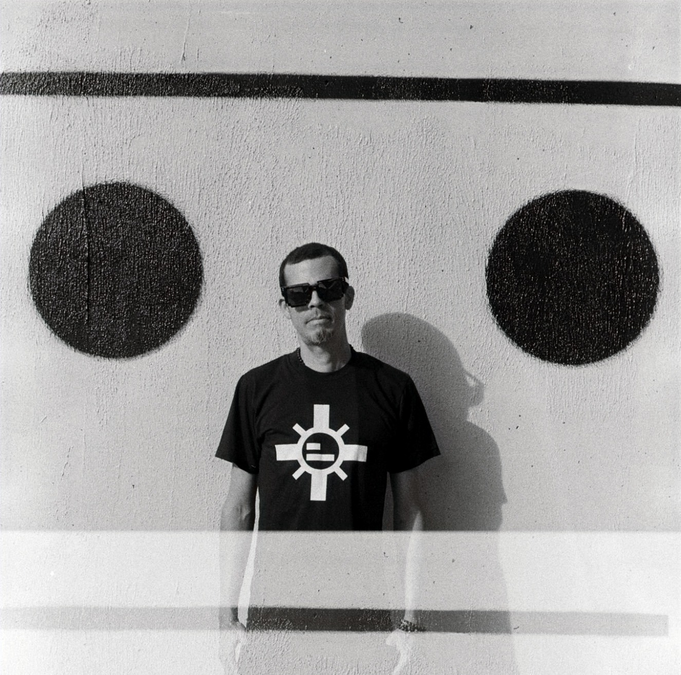 American DJ Jonnie Sparko standing in front of a wall with 2 black crcles and a line above the circles wearing sunglasses, black and white image.