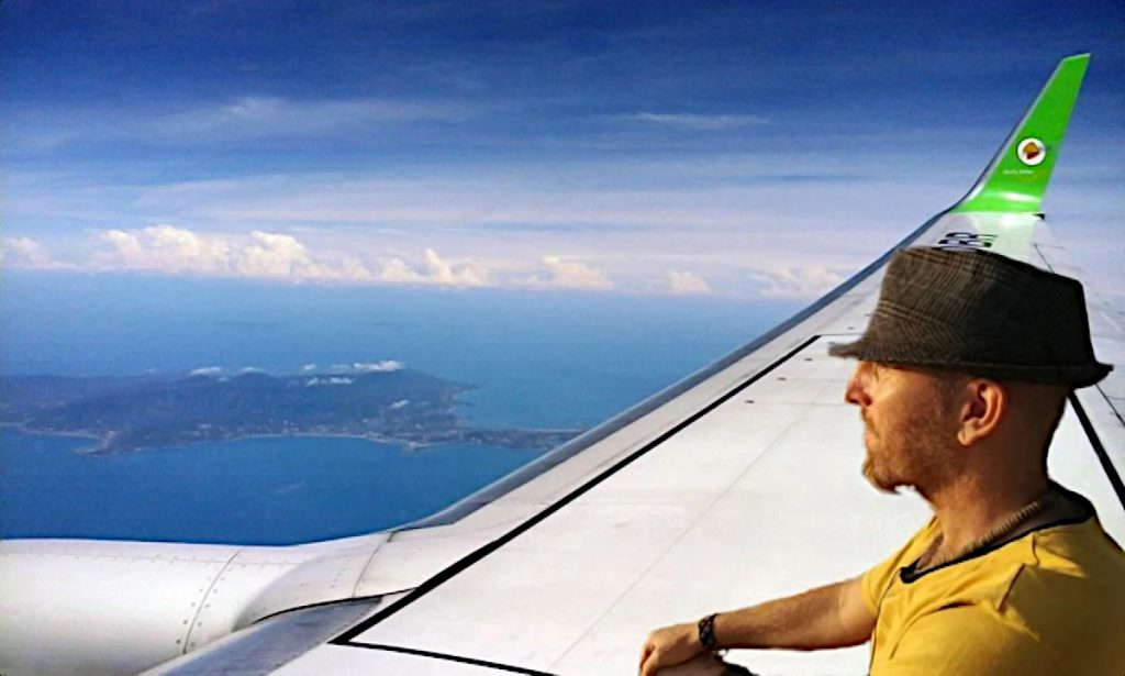 British DJ and producer Jonni Hey sitting on a wing of a commercial aircraft flying over Thailand, edited image in colour.