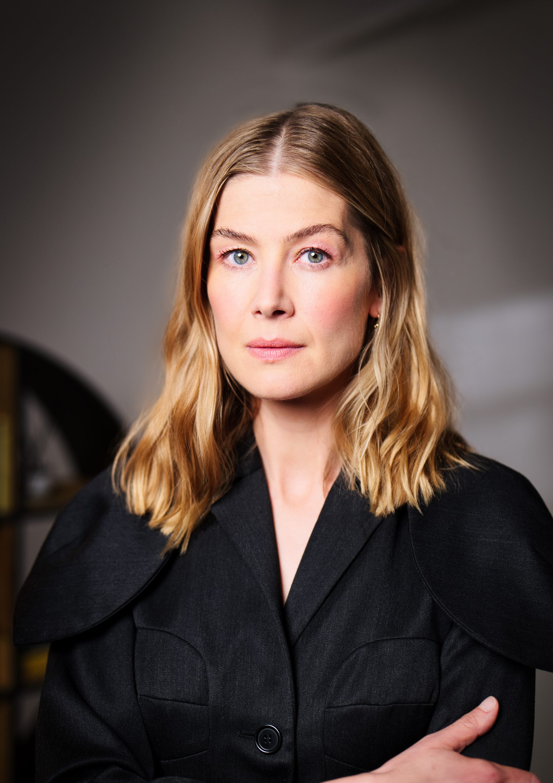 Portrait of actor Rosamund Pike wearing a dark tailored jacket, looking directly at the camera in a softly lit indoor setting. Image credit: Oliver Kingsley.