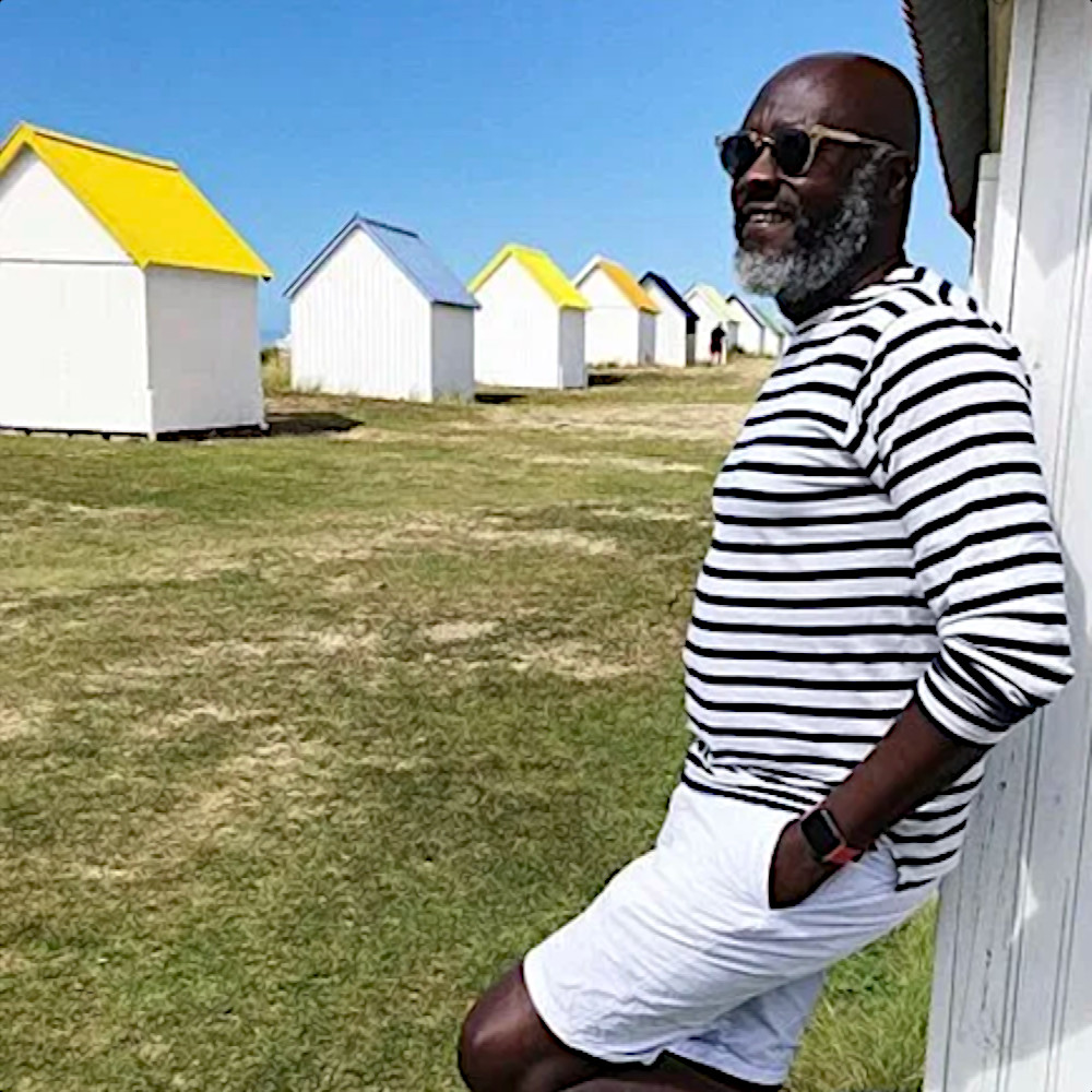 French DJ Black Sparrow leaning against a beach hut facing to his right, colour image.