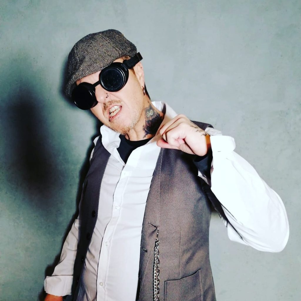 El Brujo in a flat cap and round goggles poses against a gray wall, wearing a white shirt and vest, with a bold, edgy look