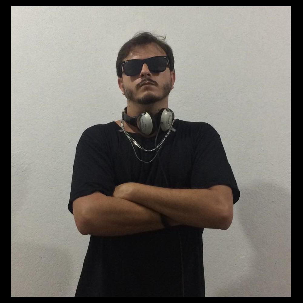 Portrait of Brazilian DJ and producer Bokard standing against a plain wall with his arms folded, wearing dark sunglasses, a black T-shirt and headphones around his neck.